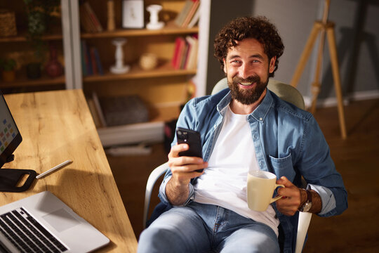 Relaxing in a comfortable chair, a man smiles while holding a coffee mug and a smartphone in a stylish home office filled with warm light and modern decor. - Powered by Adobe