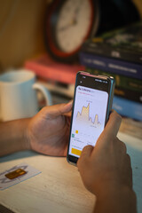 A person checks gold price charts on a smartphone while sitting at a desk with books, a clock, and a gold bar card nearby