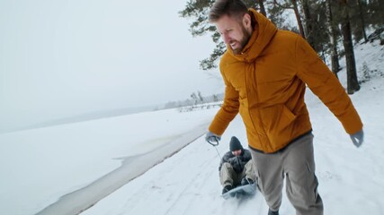 Smiling father in orange jacket pulling child on sled along snowy path near frozen lake surrounded by pine trees in cold winter day