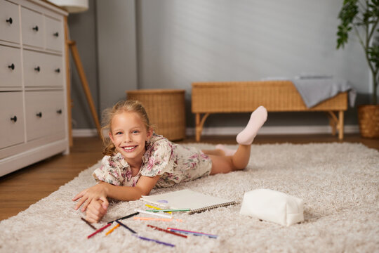 Girl smiles while lying on her stomach on a soft carpet, surrounded by colored pencils and an open sketchbook, showcasing her creativity in a bright room.