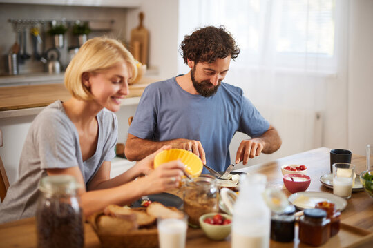 A couple is seated at a wooden dining table in a bright kitchen, happily preparing breakfast together. There are various ingredients and drinks around them, capturing a warm, intimate moment.