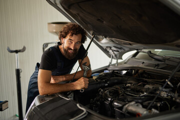 A skilled car mechanic works on a vehicle in a repair shop, checking the engine with a wrench and smiling while wearing overalls. The environment is clean and well-lit.