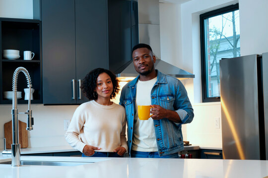 Young Black man holding yellow mug standing beside young Black woman smiling in kitchen, both looking at camera, stainless steel appliances