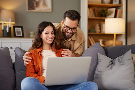 A couple sits closely on a couch, engaging with a laptop in their warm and inviting living room. The scene is relaxed, highlighting their joyful connection in the evening. - Powered by Adobe