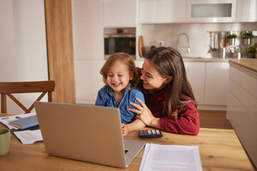A child and parent share a joyful moment at the dining table, looking at a laptop screen while surrounded by papers and a calculator. The warm environment highlights their connection.