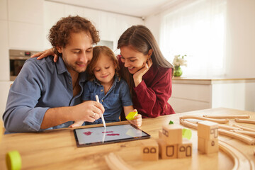 Parents and their young child share a joyful moment in the kitchen, using a tablet to draw while surrounding themselves with wooden toys and building blocks.
