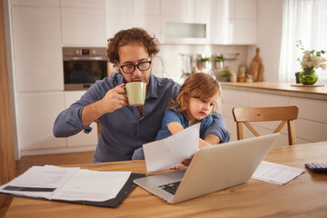 A father drinks coffee while seated at a kitchen table, focused on his laptop. His daughter explores papers next to him, enjoying quality family time in a cozy home environment.
