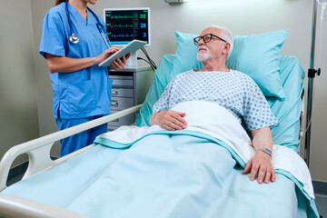 Senior man lying in hospital bed interacting with young female nurse holding digital tablet, medical monitor displaying vital signs in background