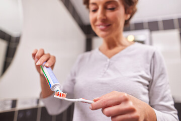 A woman is standing in a sleek bathroom, getting ready to brush her teeth. She smiles while applying toothpaste on her toothbrush, promoting good oral care habits each morning.