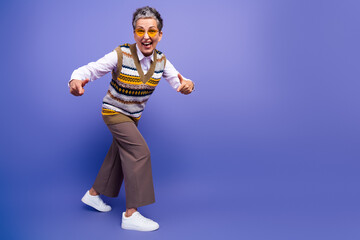 Playful mature woman with short gray hair wearing a colorful knit vest and casual shirt strikes a fun pose against a purple background