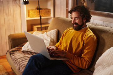 A man is focused on his laptop in a warm living room, surrounded by soft lighting and comfortable home decor. He enjoys a productive evening at home.