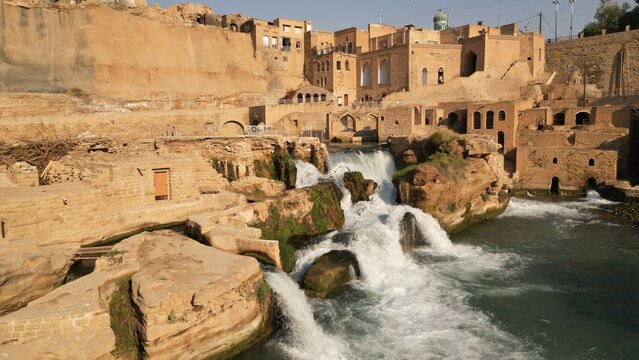 View of ancient stone buildings cascade down to meet the roaring, white waters of a waterfall, a timeless scene of human architecture, Shushtar, Khuzestan Province, Iran.