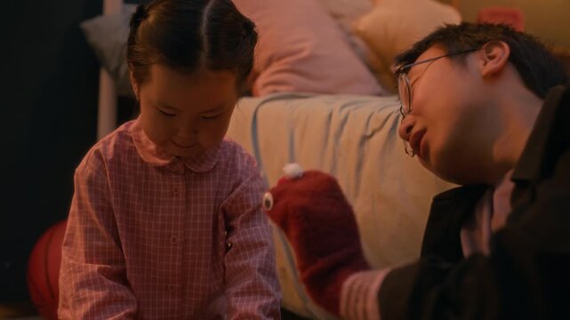 Warm evening moment of Asian father and daughter playing together at home with colorful building blocks and sock puppet in cozy, softly lit room