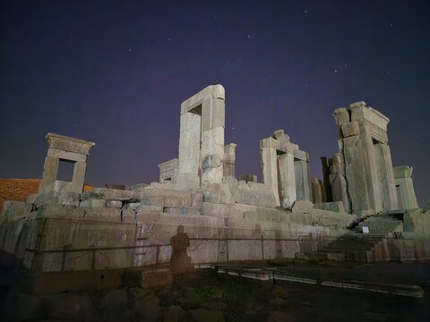 View of the ancient ruins of Persepolis under a starlit sky, shadows dancing on weathered stones, a silent story etched in time, Marvdasht, Fars Province, Iran.