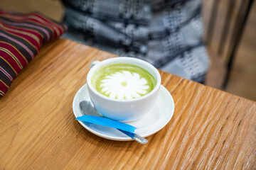 A warm cup of matcha latte rests on a white saucer atop a wooden table, with white foam forming a simple floral design in the center.