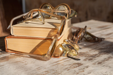 Various vintage reading glasses are scattered messily on books in bright sunlight with light reflections on the lenses on a wooden table, close-up.