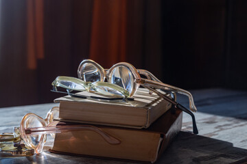 Various vintage reading glasses lie on top of books in bright evening sunlight on a wooden table, close-up.