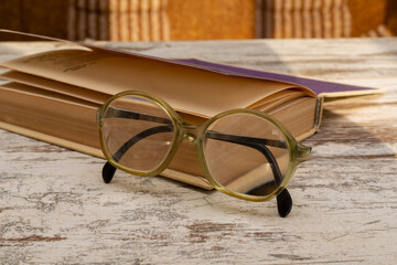  Large vintage reading glasses with strong diopters rest on a book in the evening sunlight with light reflections on the lenses on a wooden table, macro shot.