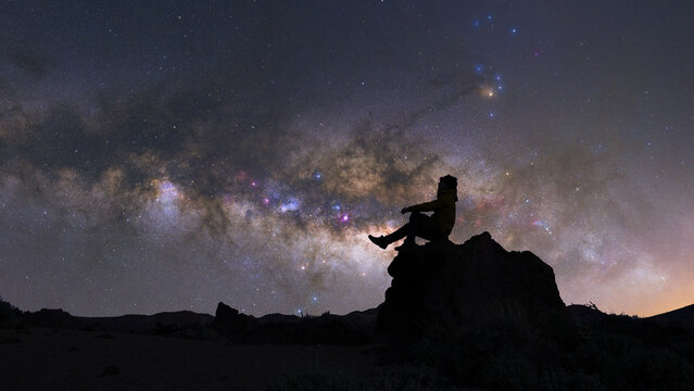 View of a lone figure silhouetted against a breathtaking, star-studded Milky Way, perched atop a rocky outcrop under the celestial canopy, Teide plateau, Tenerife Island.