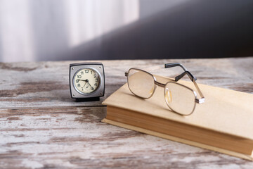 Glasses lie on a closed book next to old clocks on a rough wooden table in sunlight, close-up. Suitable for articles about vision, eye health, glasses selection, optical store advertising.