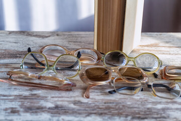 Various vintage reading glasses scattered chaotically in front of a book on a wooden table against a gray wall with light reflections from the window, close-up.