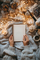Cozy winter scene with a blank notebook held over soft knits, surrounded by warm candles and glowing fairy lights
