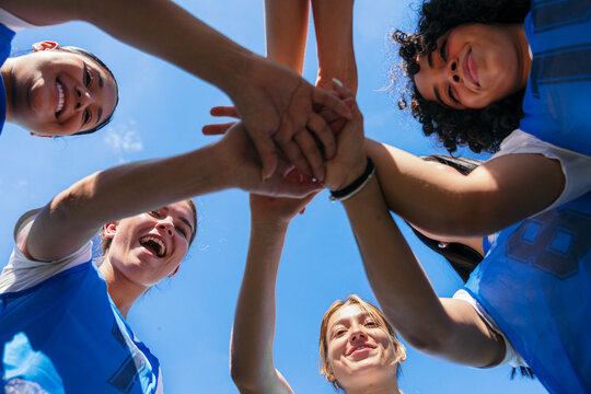 Female athletes huddling, stacking hands, and celebrating teamwork against a blue sky, showing spirit and support