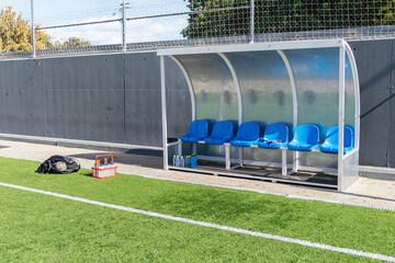 Empty coach bench on a green artificial turf football field with blue seats, water bottles, and equipment ready for training
