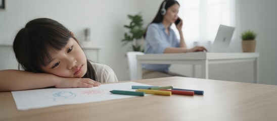 Sad little child resting head on hands and missing attention, sitting on table with crayons and drawing paper while busy mother working from home in background, handling business call