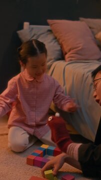 Vertical shot of Asian father and his pretty little daughter sitting on rug near cozy child bed and playing with sock puppet during evening playtime in cozy home setting