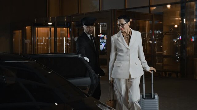 Male personal chauffeur in uniform opening car door for businesswoman then helping carrying suitcase while meeting high profile client at hotel entrance in evening