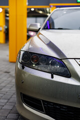 Fototapeta premium Shiny silver car with rain droplets on the headlight, captured in a close-up shot at a brightly lit car wash station