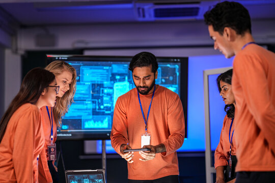 In a government command center, a diverse team of intelligence analysts collaborates on a critical mission, analyzing real-time data from a tablet, phone, and a large video wall. - Powered by Adobe