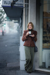 A woman enjoys a warm drink outside a shop on a foggy street, dressed in a textured brown jacket and green trousers, calmly leaning against the doorway.