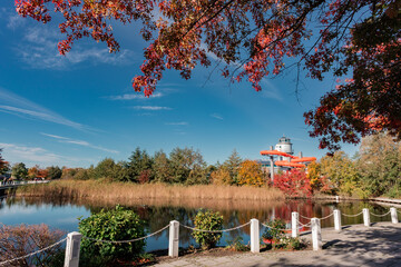 View across a calm pond to a spa/leisure center with orange water slides, framed by vibrant red and gold autumn foliage and a clear blue sky.