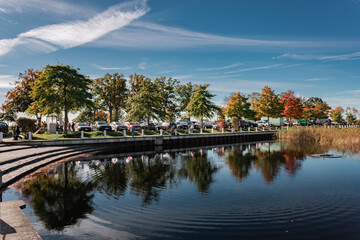 Paved autumn pathway along a pond or small lake, lined with trees and a bollard chain fence. Parked cars are visible on the left, with two people enjoying the view.