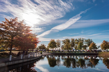 Paved autumn pathway along a pond or small lake, lined with trees and a bollard chain fence. Parked...