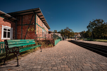 Historic narrow-gauge railway station platform with a traditional brick and timber-framed building. A person stands waiting, next to the tracks under a clear blue sky.