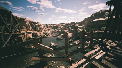 A weathered wooden bridge spans a sparkling river surrounded by rugged rocks and hills. Fluffy clouds drift across the bright sky, creating a serene atmosphere. © icetray