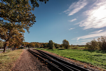 Perspective view of the narrow-gauge railway tracks leading into the distance, lined by trees with early autumn foliage under a dramatic blue sky with wispy clouds.