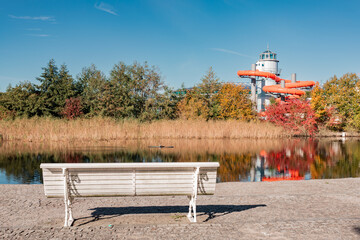 A white bench overlooks a pond reflecting autumn trees and the tower and orange water slides of a modern spa or leisure facility in the background.