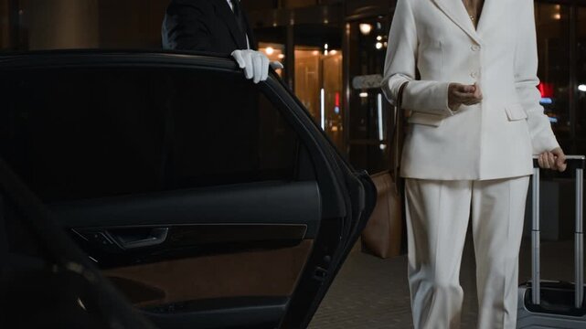 Male personal driver in white gloves opening car door for businesswoman helping important client with loading baggage into vehicle at hotel entrance in evening