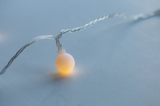 Warm glowing LED string light on a soft pastel surface. Minimalistic, cozy aesthetic with a focus on a single illuminated bulb and twisted transparent wire. - Powered by Adobe