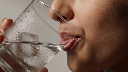 Extreme close up of a young woman's lips drinking refreshing cold water with ice cubes from a transparent glass, quenching her thirst and smiling with satisfaction in a studio setting