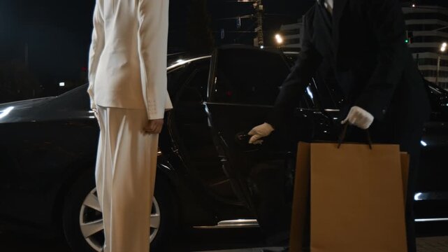 Middle aged woman in white suit getting into back seat of luxurious car while male personal chauffeur assisting VIP passenger with opening door and loading shopping bags into vehicle in night city
