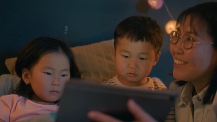 Asian mother and two young children sitting in bed using tablet together at night, probably reading or watching story before going to sleep - Powered by Adobe
