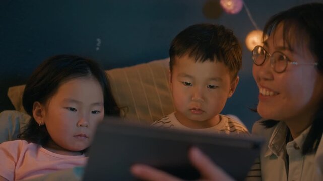 Asian mother and two young children sitting in bed using tablet together at night, probably reading or watching story before going to sleep