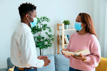 Black man and Black woman standing indoors wearing medical masks, engaging in conversation while woman holding open notebook, both appearing as young adults in modern setting