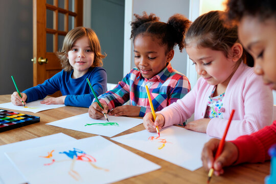 Group of diverse children drawing with colored pencils at table, Caucasian boy, Black girl, two Caucasian girls focused on creative activity, watercolor set and drawings visible