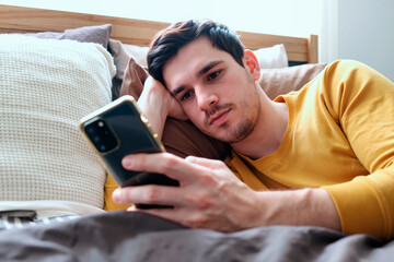 Portrait of Caucasian young adult man lying on bed holding smartphone in hand looking at screen with serious expression, relaxing in bedroom during daytime